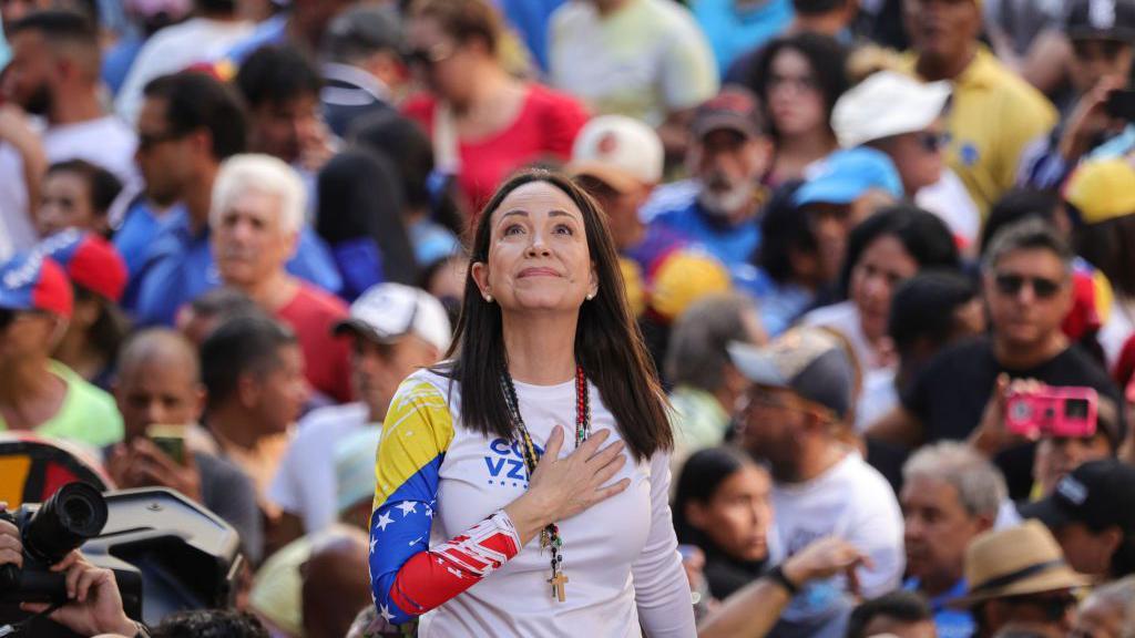 María Corina Machado de pie, con una mano en el pecho, tiene la vista al cielo. Viste una camisa con los colores de la bandera venezolana, azul, amarillo y rojo. También tiene blanco. Detrás de ella se ve una multitud de gente difuminada. 