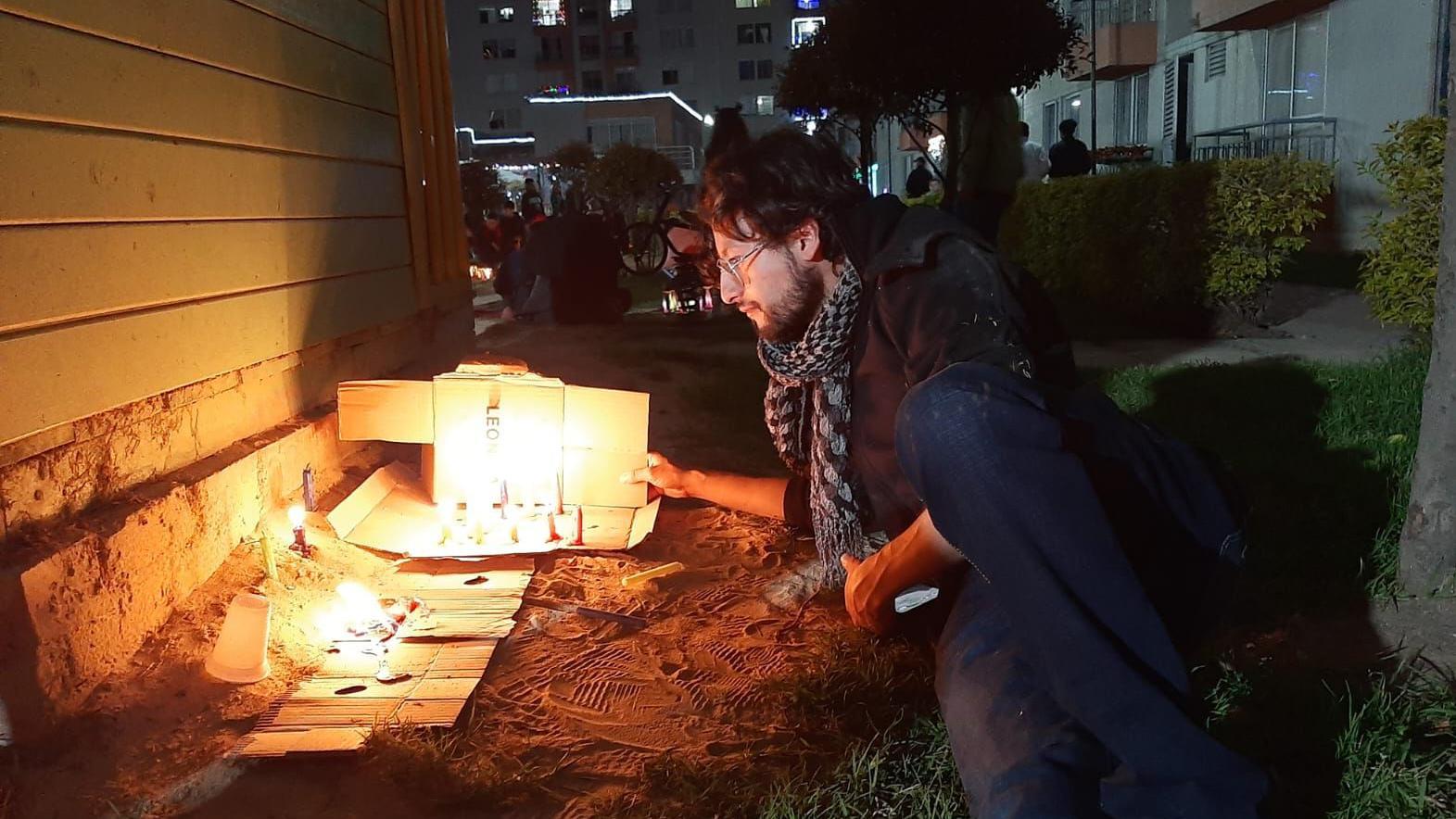 Manuel Tique, tumbado sobre el césped durante la noche frente a lo que parece ser un pequeño memorial con flores y velas.