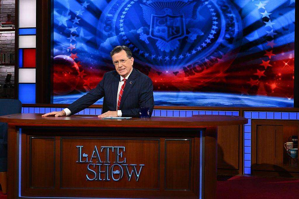 Stephen Colbert sentado detrás del escritorio de “Late Show”, con un fondo rojo y azul con estrellas.