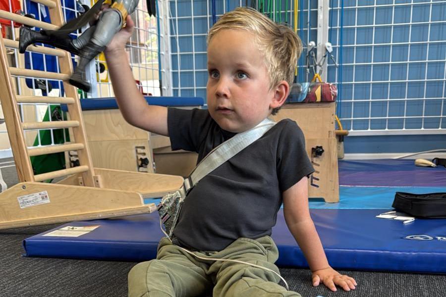 Edward está sentado en el piso sosteniendo un muñeco de juguete. Viste una playera oscura y pantalón de sudadera verde 