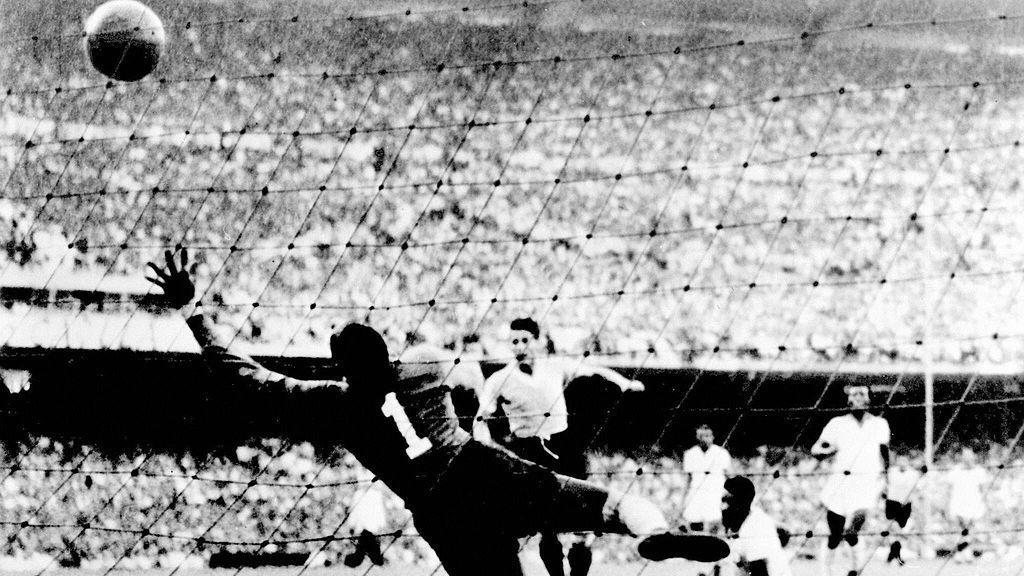 A imagem em preto e branco mostra um lance de gol em uma partida de futebol, com a bola j&aacute; ultrapassando o alcance do goleiro, que salta em v&atilde;o em dire&ccedil;&atilde;o ao canto superior da meta. Ao fundo, jogadores acompanham a jogada diante de um est&aacute;dio lotado, em um registro hist&oacute;rico que destaca o momento decisivo da final.