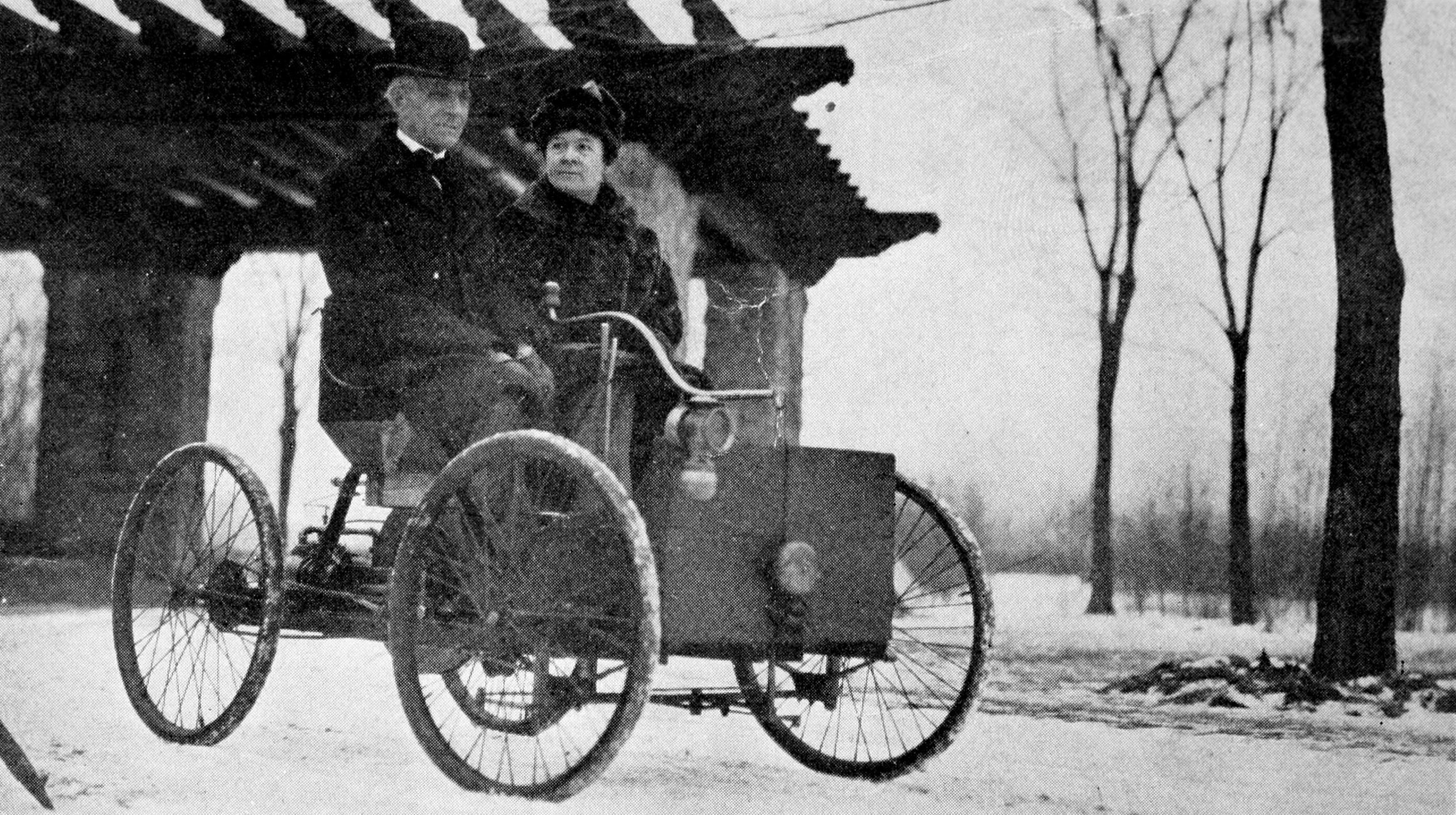 A imagem em preto e branco mostra duas pessoas sentadas em um ve&iacute;culo antigo de quatro rodas finas e estrutura simples, semelhante aos primeiros autom&oacute;veis. O homem conduz o ve&iacute;culo enquanto a mulher est&aacute; sentada ao lado dele, ambos usando roupas pesadas e chap&eacute;us t&iacute;picos do in&iacute;cio do s&eacute;culo 20. Ao fundo, aparecem &aacute;rvores sem folhas e uma constru&ccedil;&atilde;o com telhado inclinado em uma paisagem de inverno.