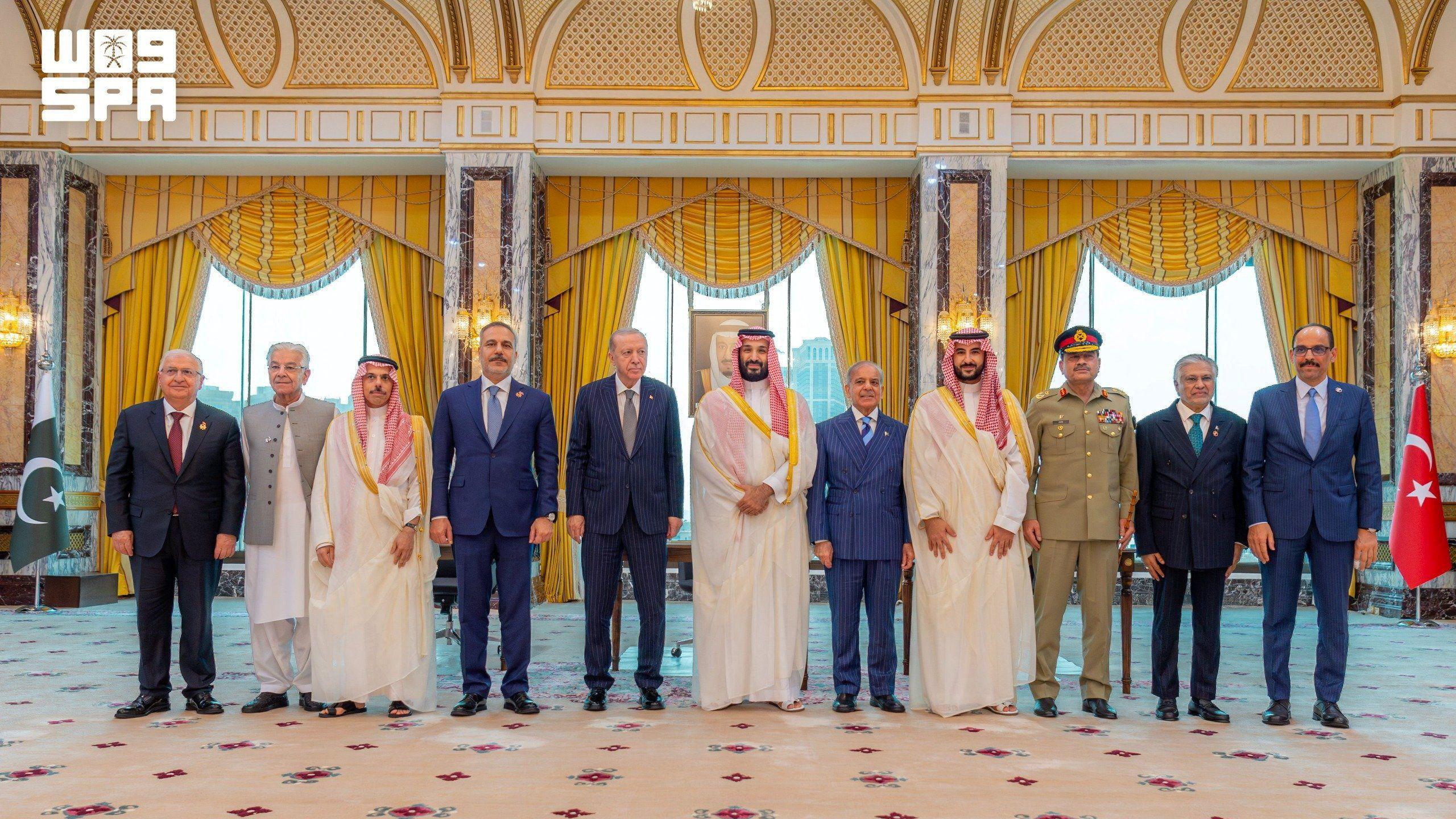 Líderes y figuras políticas de Turquía, Arabia Saudita y Pakistán posan para una foto. Visten una mezcla de túnicas blancas sauditas, trajes occidentales, uniformes militares y túnicas pakistaníes. Cortinas a rayas amarillas cuelgan de las ventanas detrás de ellos. 