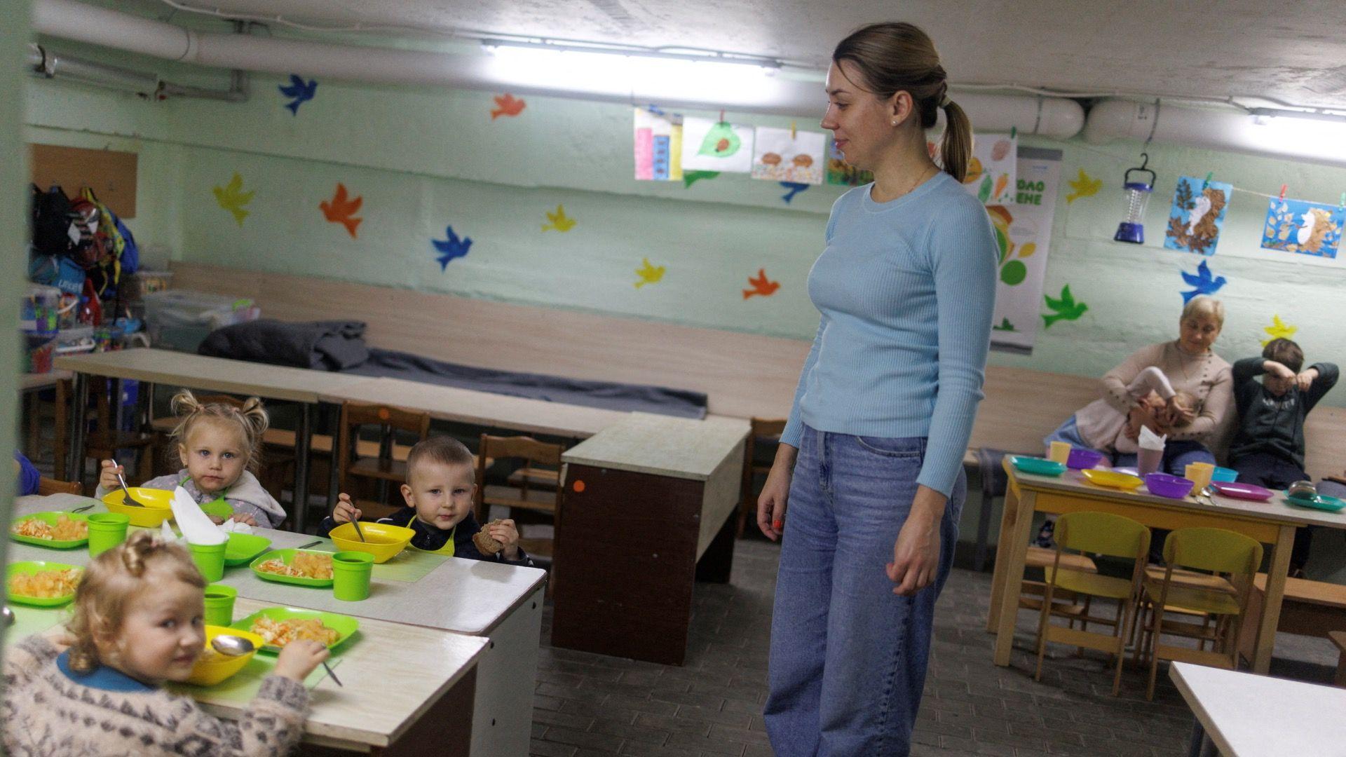 Maryna Ponomarenko, de 40 años, empleada de un jardín de infancia, sonríe mientras observa a los niños sentados a una mesa comiendo en cuencos, el 10 de octubre de 2025. Se encuentran dentro de un refugio antibombas y la cabeza de la Sra. Ponomarenko está cerca del techo. Recortes de papel multicolor con forma de pájaros adornan las paredes verde claro. Al fondo, una mujer está sentada con una niña recostada sobre su regazo y otro niño sentado a su izquierda, tirando de los cordones de sus pantalones. Dibujos hechos a mano cuelgan de pinzas sujetas a las tuberías.
