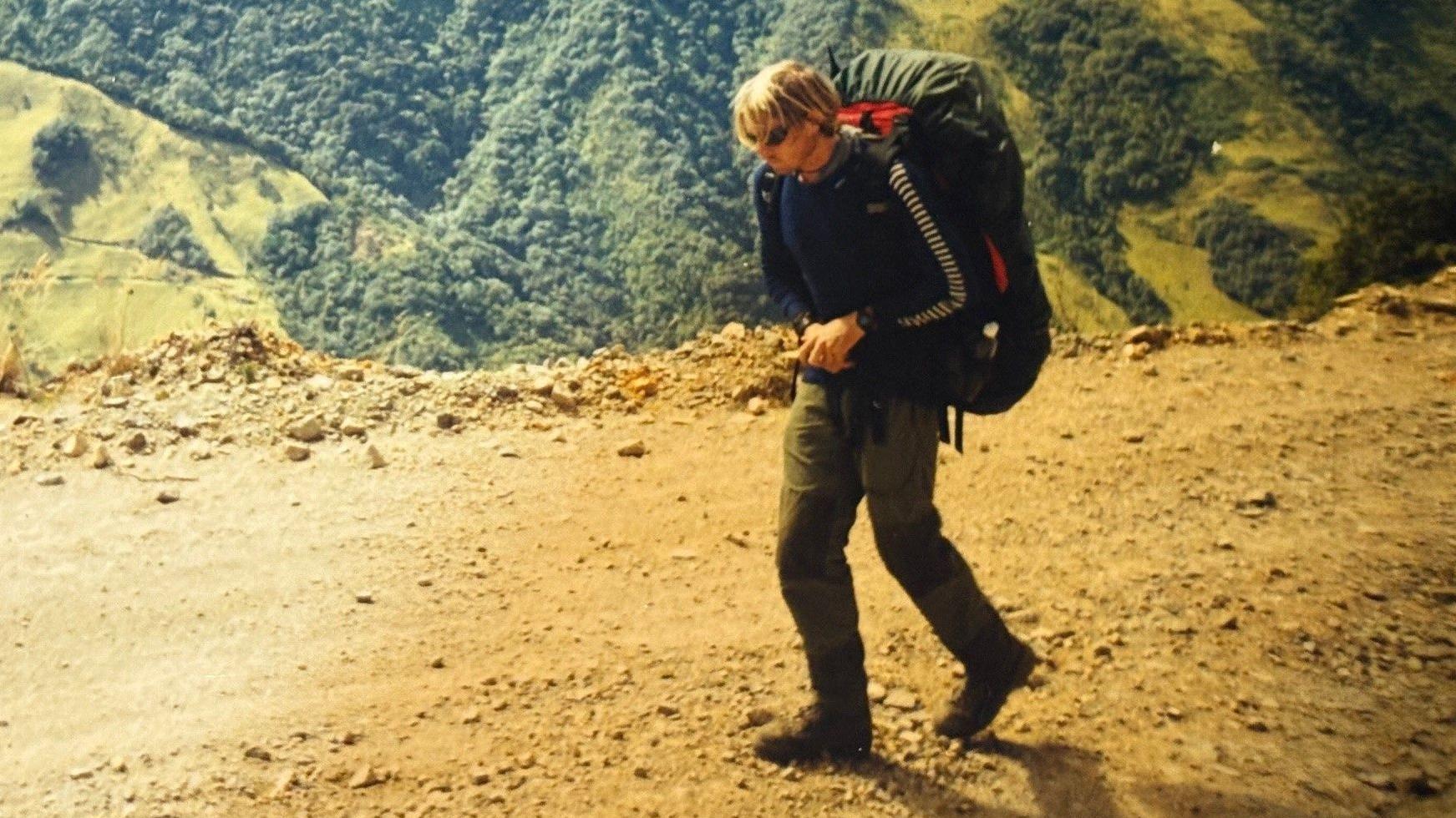 Um homem de cabelos loiros, com barba por fazer, caminha por uma montanha, com uma floresta densa abaixo. Ele carrega uma grande mochila, coberta por uma capa verde