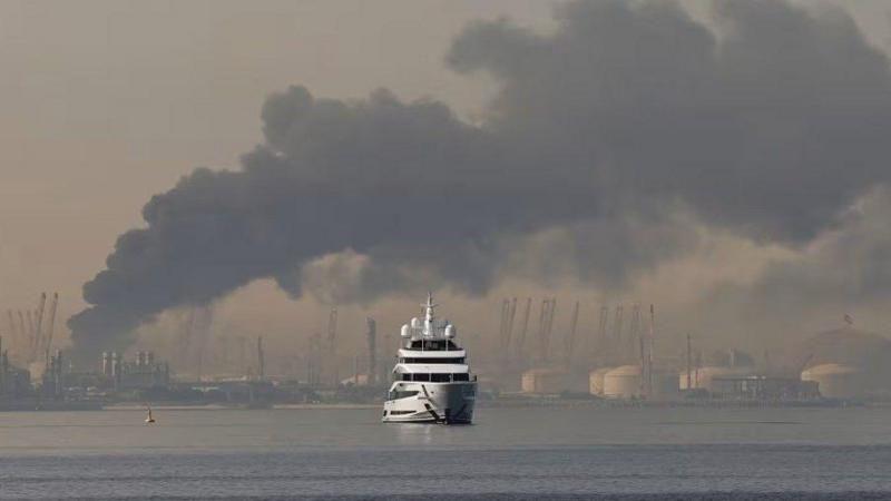 يخت فاخر في الخليج بينما يتصاعد الدخان في الخلفية من منطقة جبل علي الصناعية في الإمارات وتظهر خزانات النفط العملاقة في الخلفية