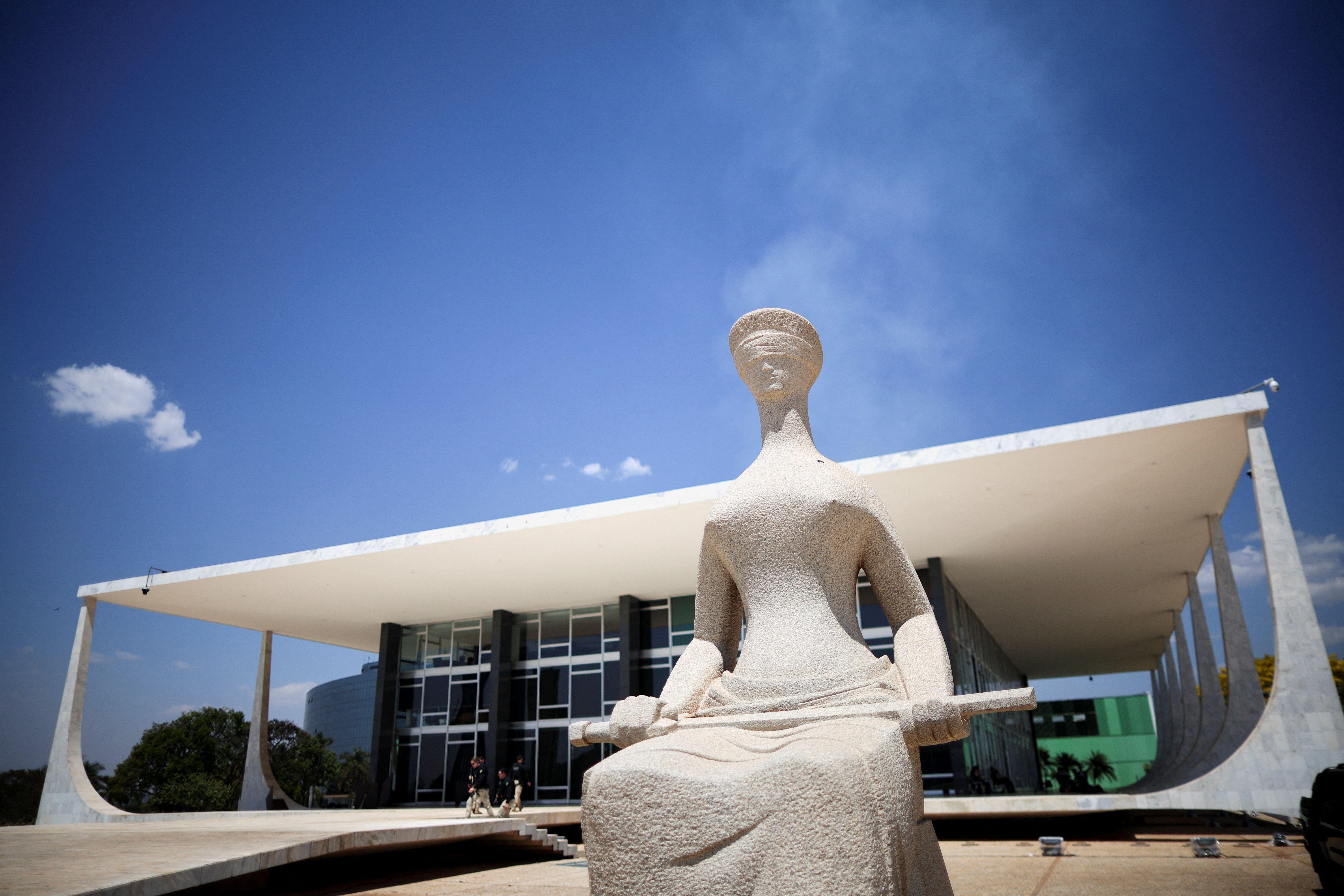 Estátua de pedra da Justiça em primeiro plano, sentada e segurando uma espada, em frente ao prédio do Supremo Tribunal Federal, em Brasília, sob céu azul intenso.