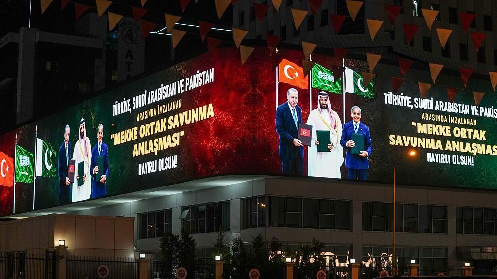 Imagem noturna com uma tela digital mostrando Erdogan, Bin Salman e Sharif mostrando documentos relativos ao acordo, com as bandeiras dos seus pa&iacute;ses ao fundo. O texto em turco ao lado deles diz: 