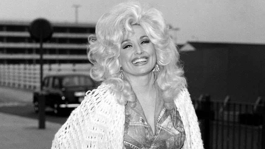 Dolly Parton sonriendo a la cámara con sus rizos dorados, un chal blanco y un vestido de jean.