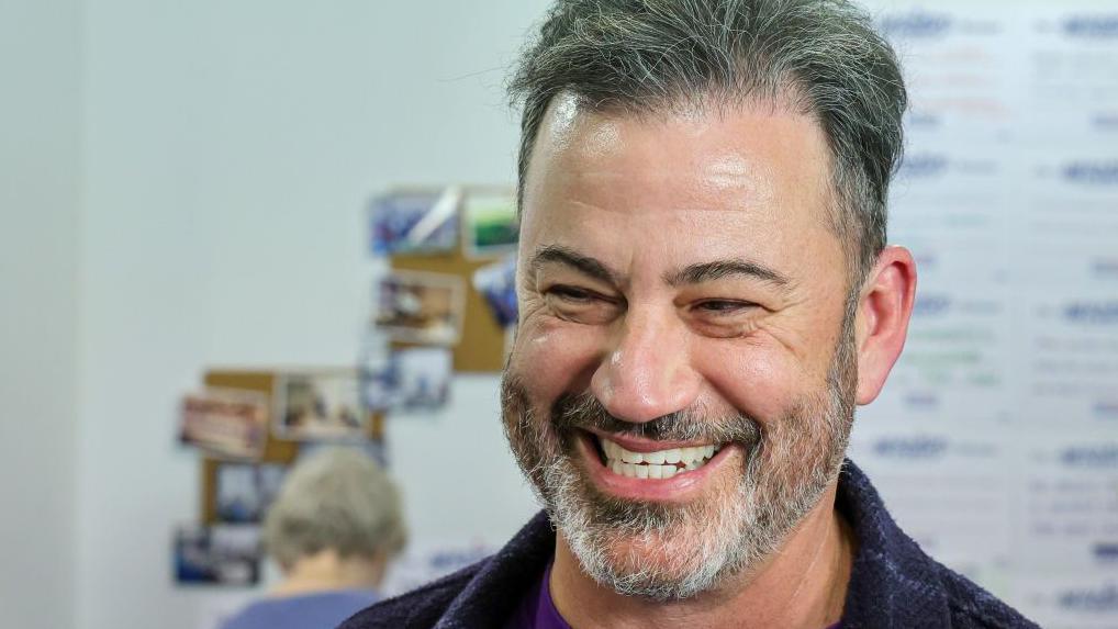 Jimmy Kimmel, sonriente, ante un fondo difuminado en el que se ve la parte trasera de la cabeza de una persona