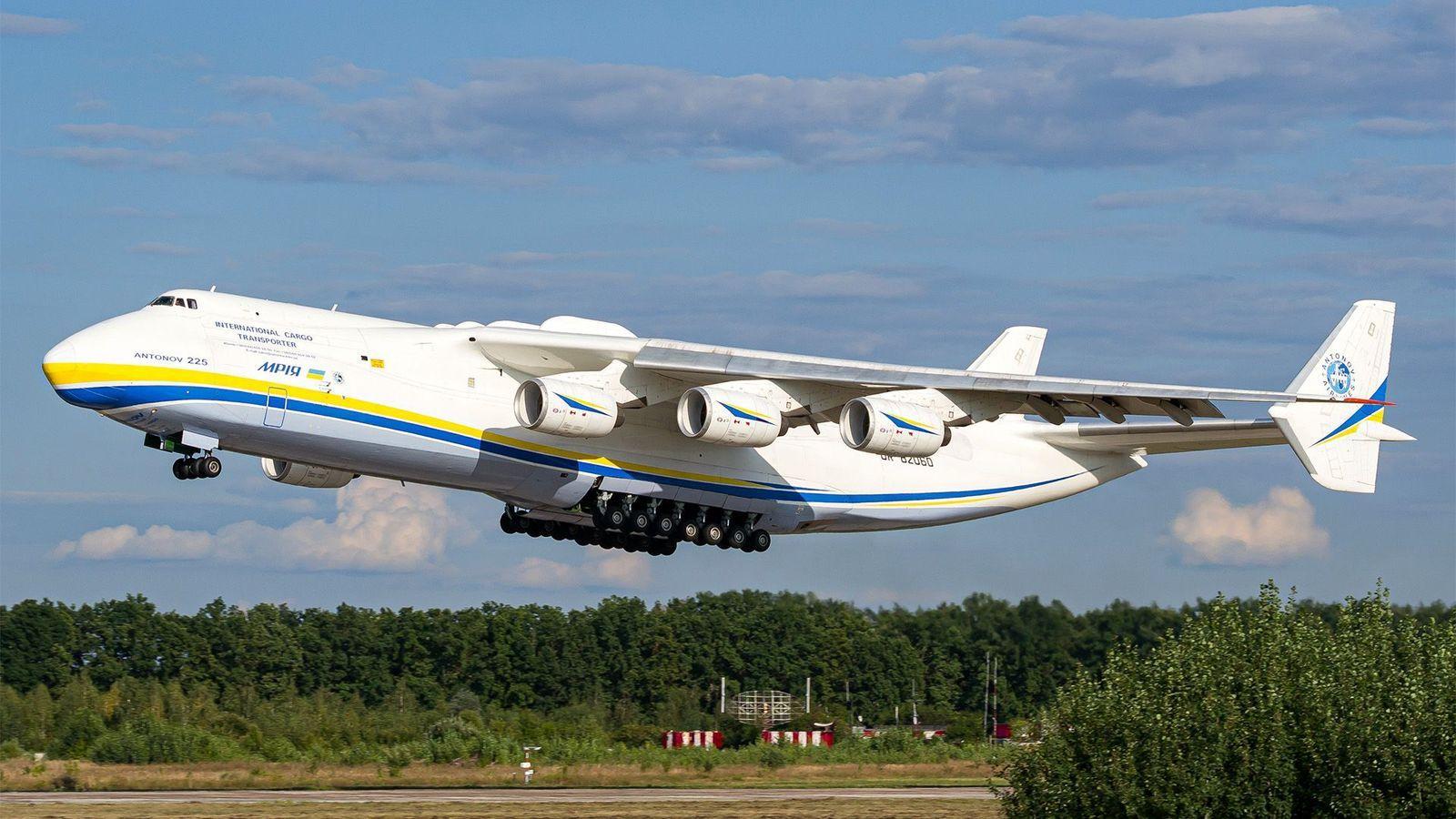 Un gran avión blanco adornado con rayas amarilla y azul a lo largo del fuselaje despega de una pista bordeada de árboles, bajo un cielo azul con unas nubes blancas. El avión tiene seis turbinas y un enorme tren de aterrizaje de 14 ruedas en el medio