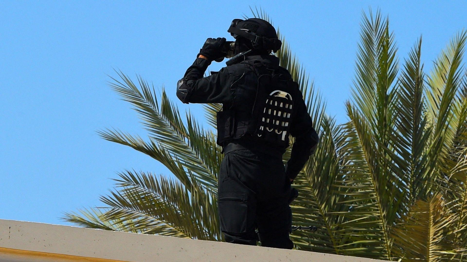 Un guardia de seguridad bahreiní se encuentra sobre una azotea junto a una palmera en un día soleado. Se lleva la mano al rostro y viste un uniforme negro con casco negro.