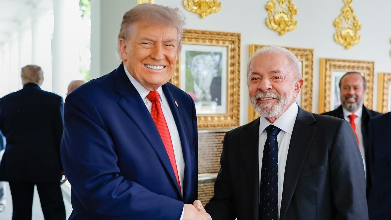 O presidente dos Estados Unidos, Donald Trump, aperta a m&atilde;o do presidente brasileiro Luiz In&aacute;cio Lula da Silva durante um encontro na Casa Branca. Ambos sorriem para a c&acirc;mera em um ambiente interno, com quadros dourados e outras pessoas desfocadas ao fundo.