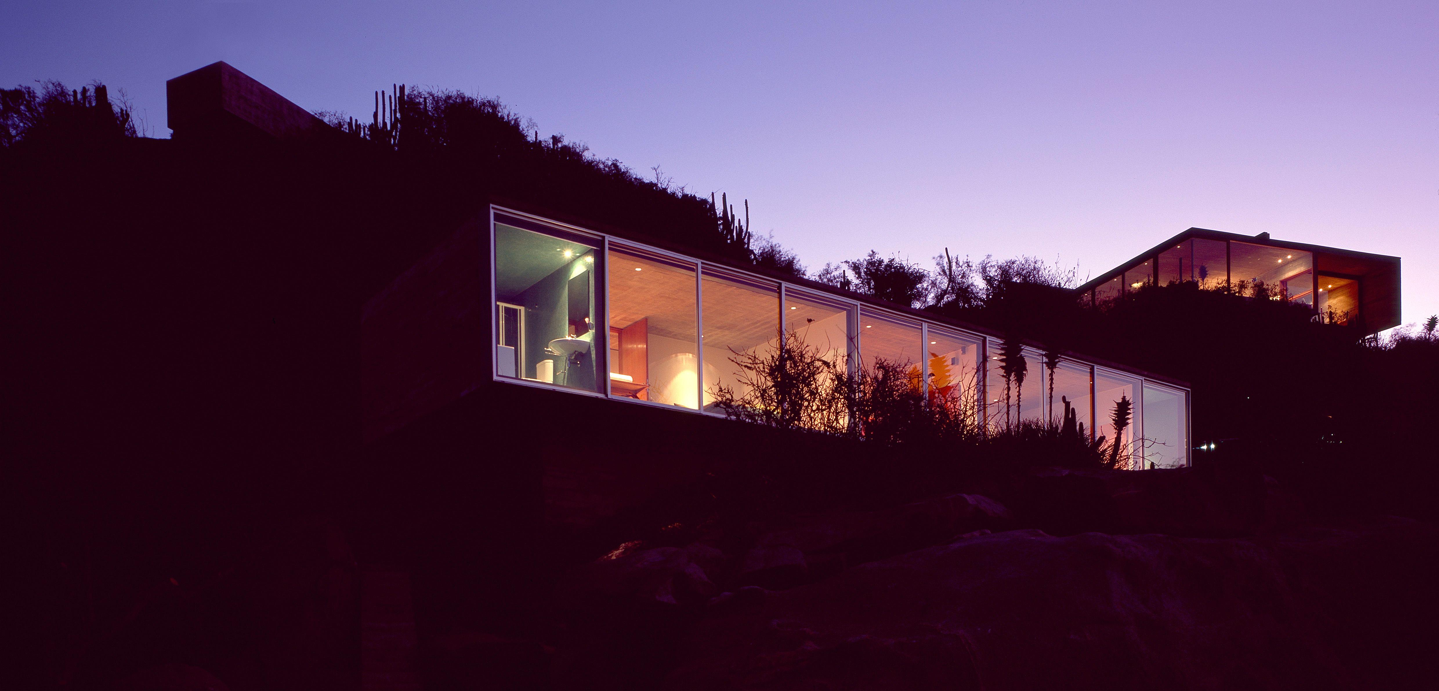 Casa moderna acristalada incrustada en una ladera, iluminada desde el interior al anochecer con un cielo violeta de fondo.