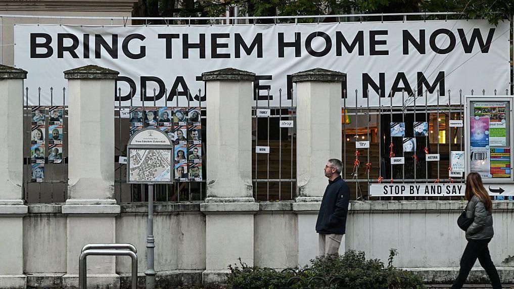 Dos personas caminan delante de un edificio en el que un gran cartel dice Bring Them Home ("tráiganlos a casa"), pidiendo el retorno de los rehenes que están en manos de Hamás