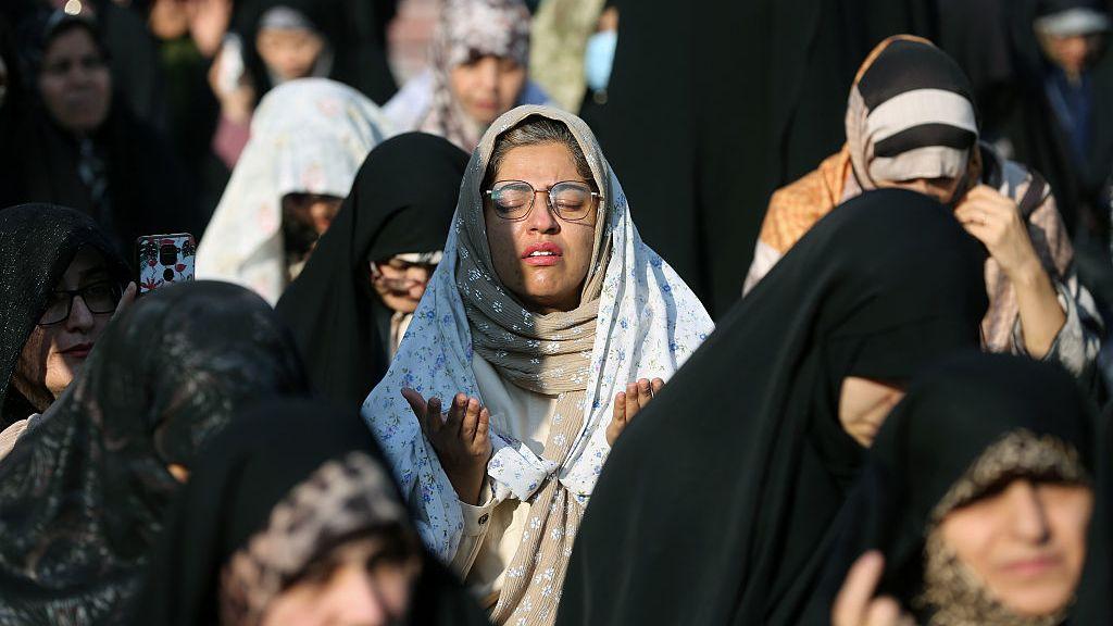 可見多名戴著面紗的婦女在德黑蘭祈禱。大多數人身穿黑色頭巾、低頭祈願；畫面中央一名戴眼鏡的女子則仰起臉、閉上雙眼，雙手合十祈禱