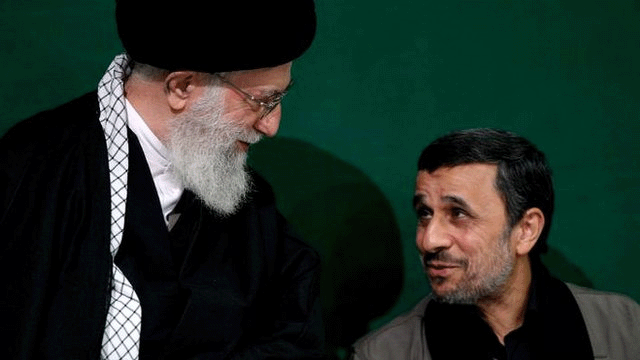Khamenei looking downwards at Ahmadinejad, who is talking and appearing to be partially smiling. Khamenei is wearing a traditional black robe and black head covering. Ahmadinejad has a black open-necked shirt, a light grey jacket and no head covering. He has a thin grey beard and black hair that is partially grey.