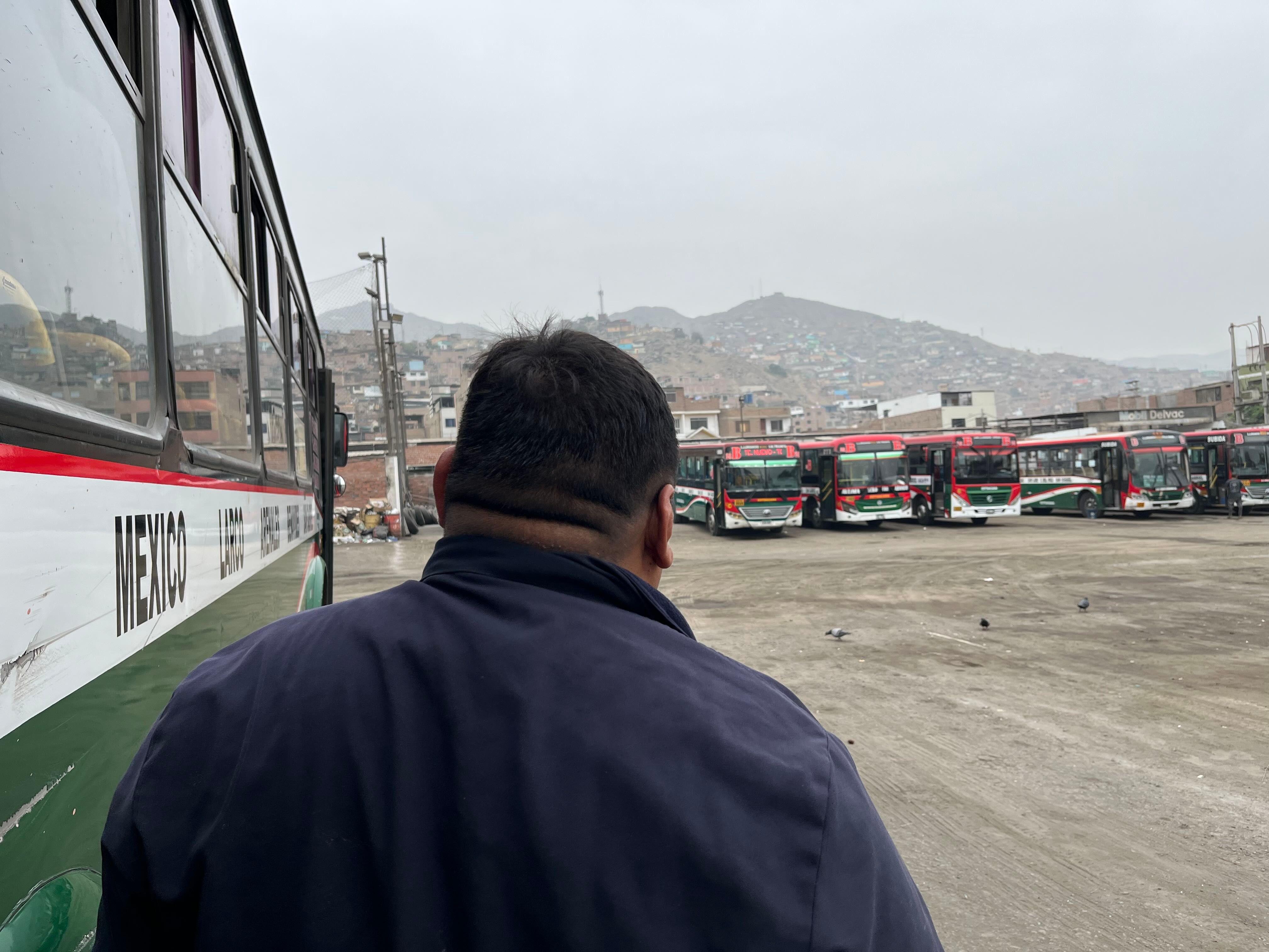 Toño, un hombre con una chaqueta azul, está parado al lado de un bus en un gran lote. Hay más buses al fondo del lote y detrás, se ve una montaña con barrios de invasión.