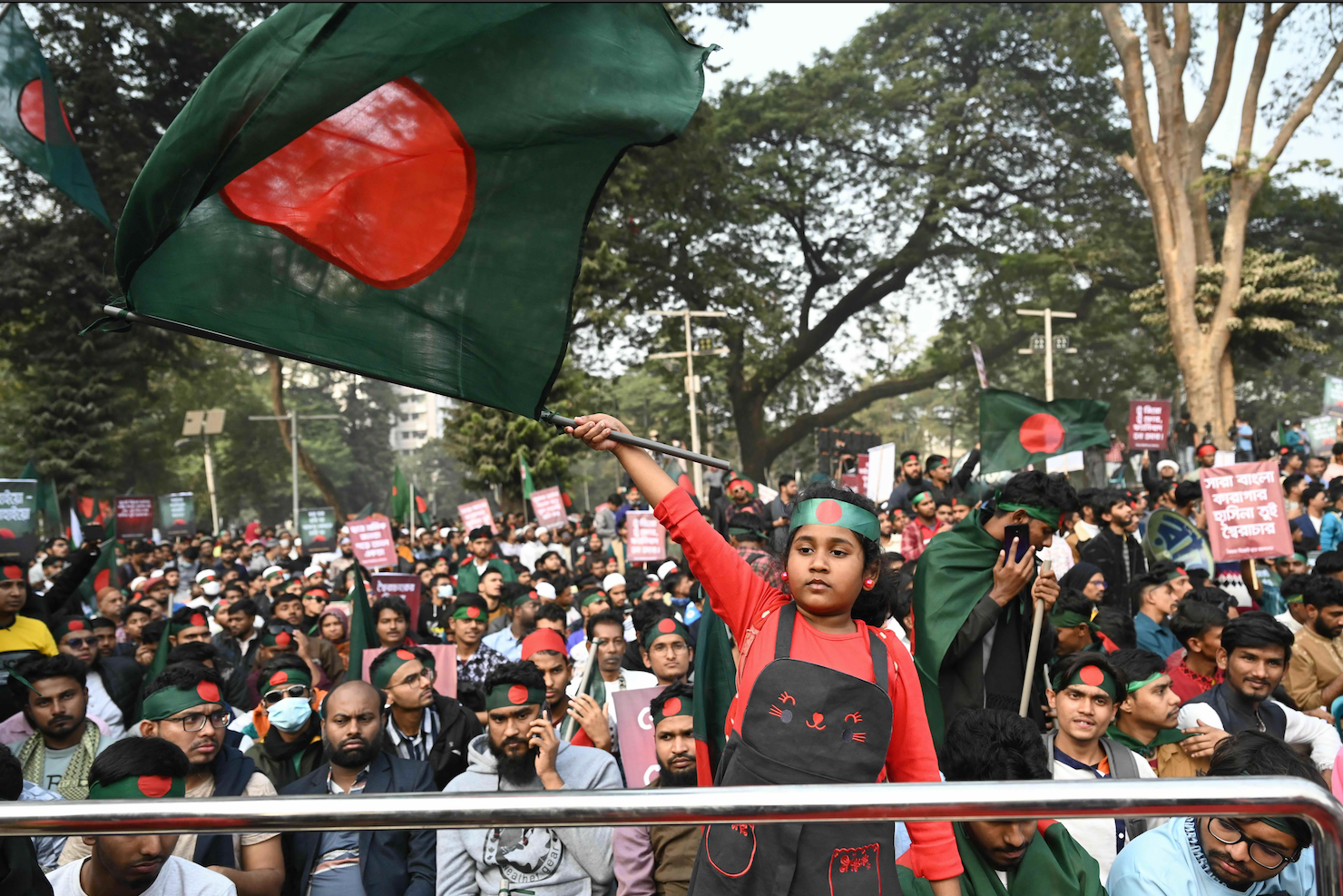 Uma multidão na rua usando faixas na cabeça com a bandeira de Banglsdesh, verde e tem um círculo vermelho.