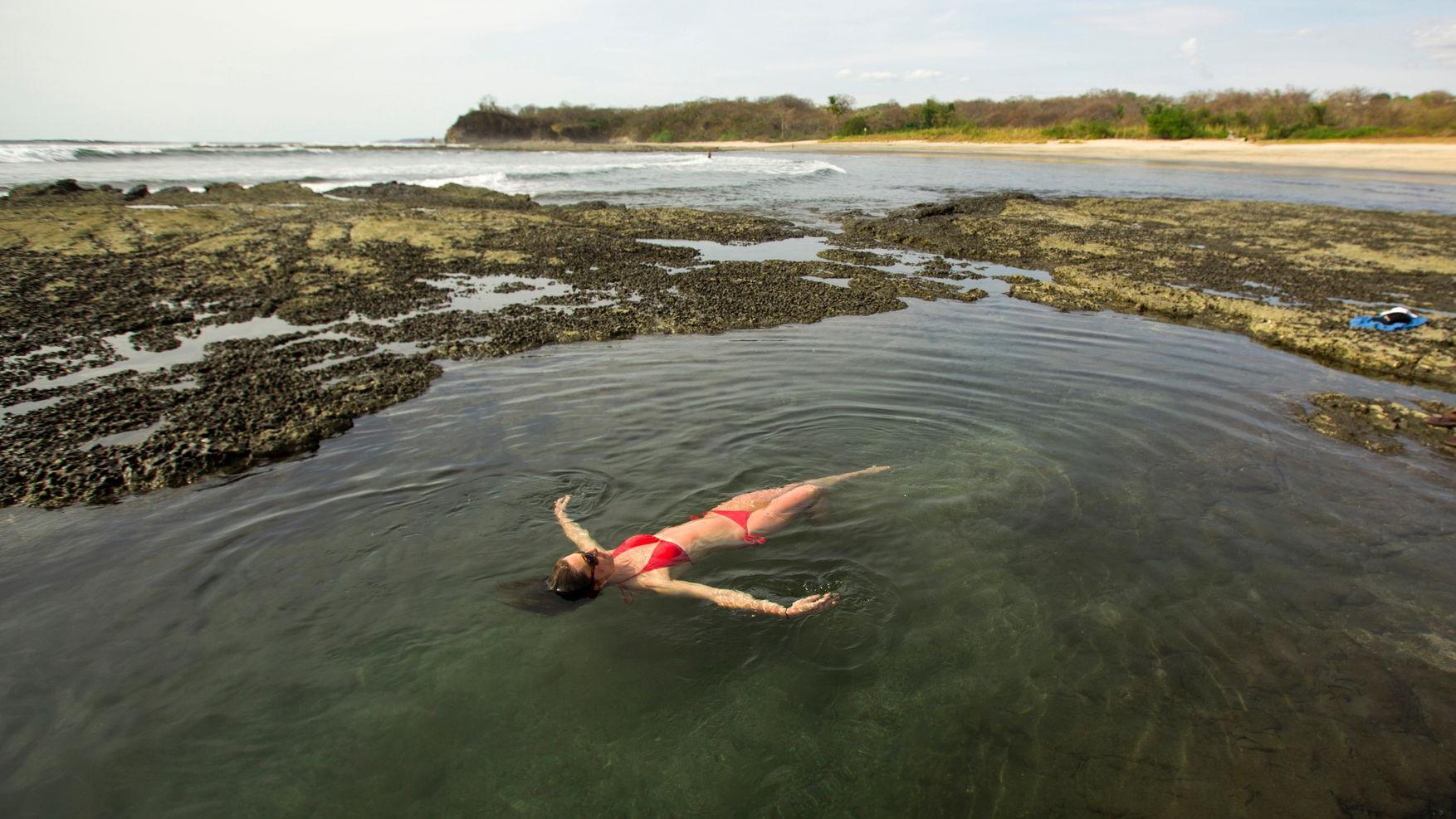 Una mujer con bikini rojo se relaja en una piscina natural. Al fondo se ve la playa.