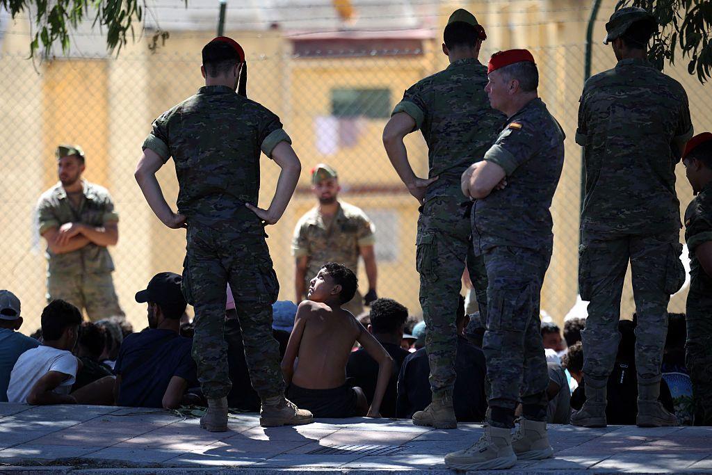 Un grupo de personas sentadas en el suelo al aire libre. A su alrededor un grupo militares españoles de pie los vigila. Entre las personas sentadas, un niño se gira y eleva la mirada hacia uno de los militares. 