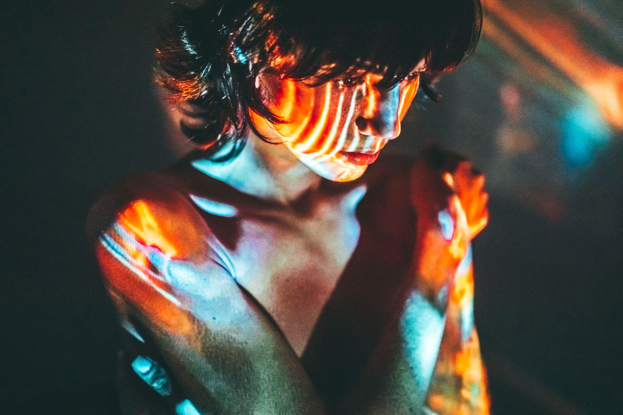 Mujer pensativa en una habitación oscura y con su cuerpo iluminado por imágenes de colores.