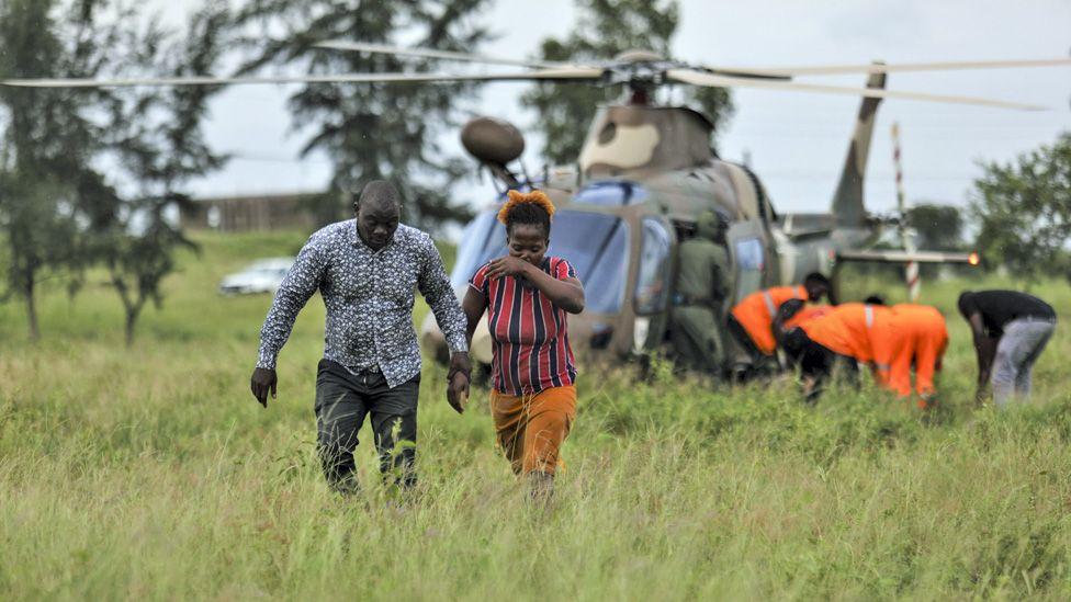 Um homem e uma mulher afastam-se de um helic&oacute;ptero militar enquanto membros de uma equipe de resgate com casacos laranja ajudam algu&eacute;m numa maca em Mo&ccedil;ambique