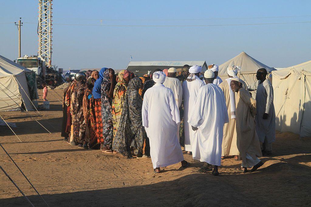 من الخلف يظهر عدد من النازحين الرجال والنساء يحتشدون قرب خيام نزوح بيضاء. إحدى النساء يظهر في يدها وعاء معدني للطعام. الأرض رملية. السماء زرقاء صافية.