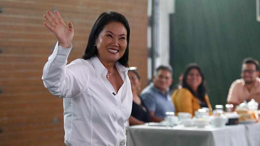 Keiko Fujimori saluda con la mano sonriente. Un grupo de personas la observa desde una mesa. 