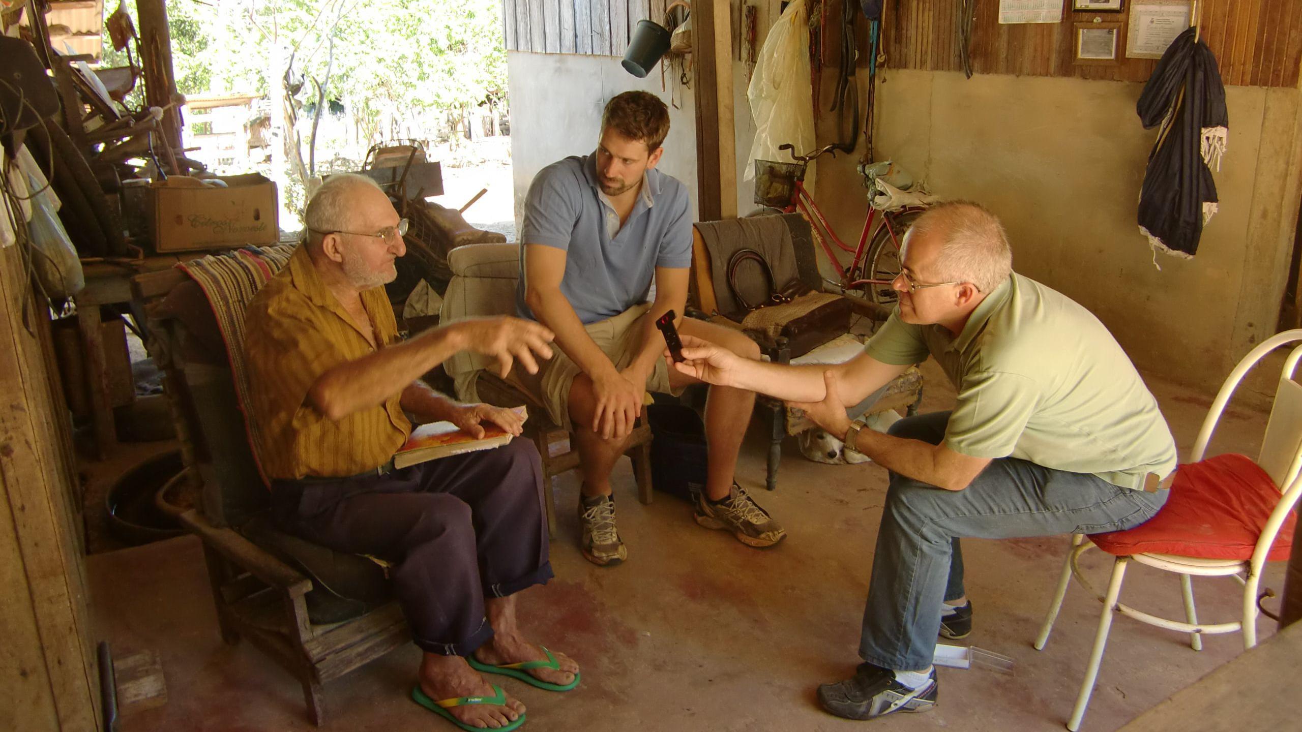 Três homens conversando sentados em ambiente doméstico; Cleo segura um gravador