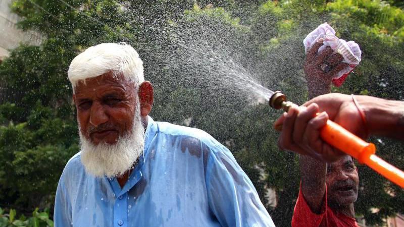 يد تمسك بخرطوم لرش المياه على وجه رجل عجوز بلحية بيضاء وبجوراه شخص يبلل قطعة قماش بالماء 