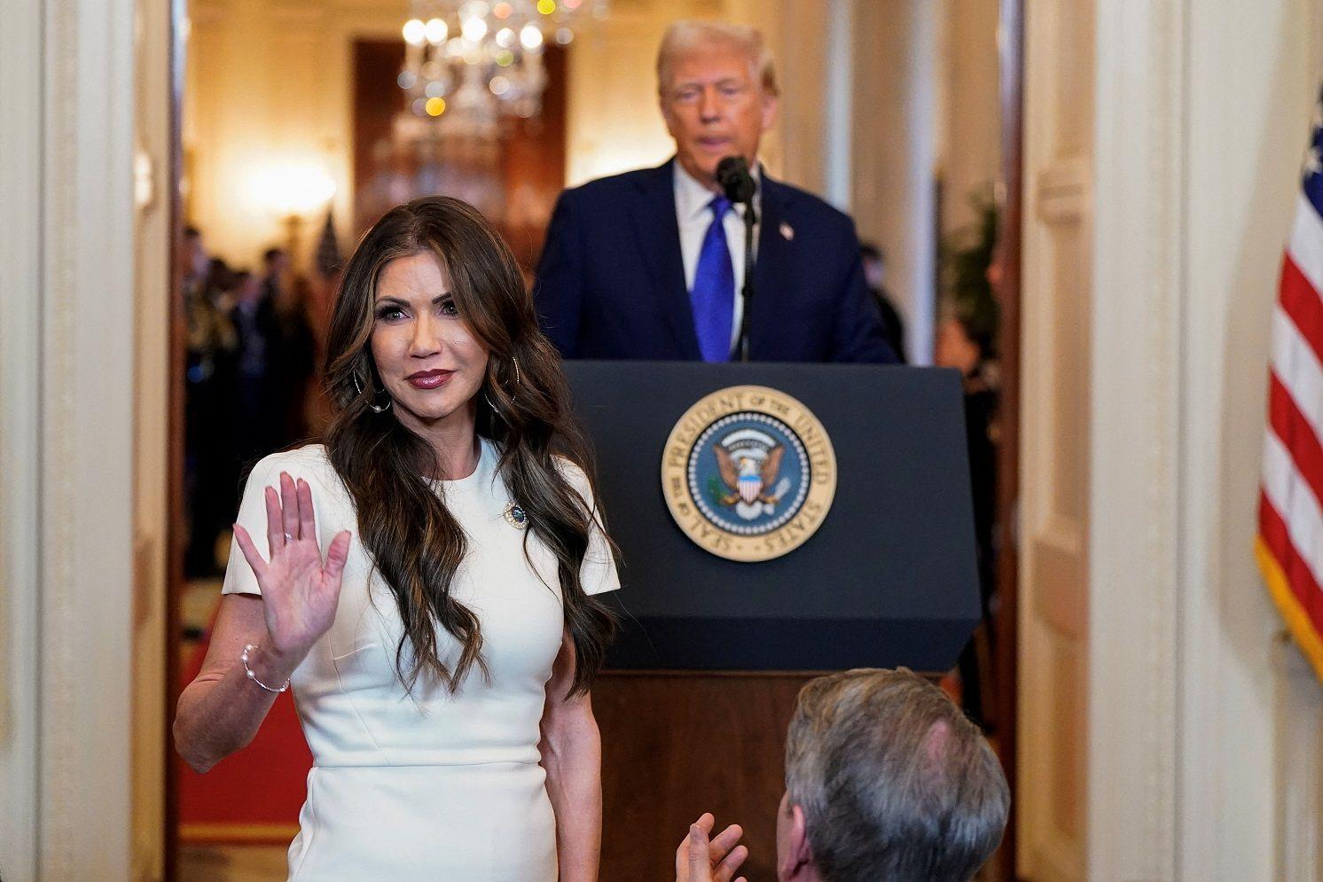 Mulher em pé acenando com a mão; atrás, Trump em palanque