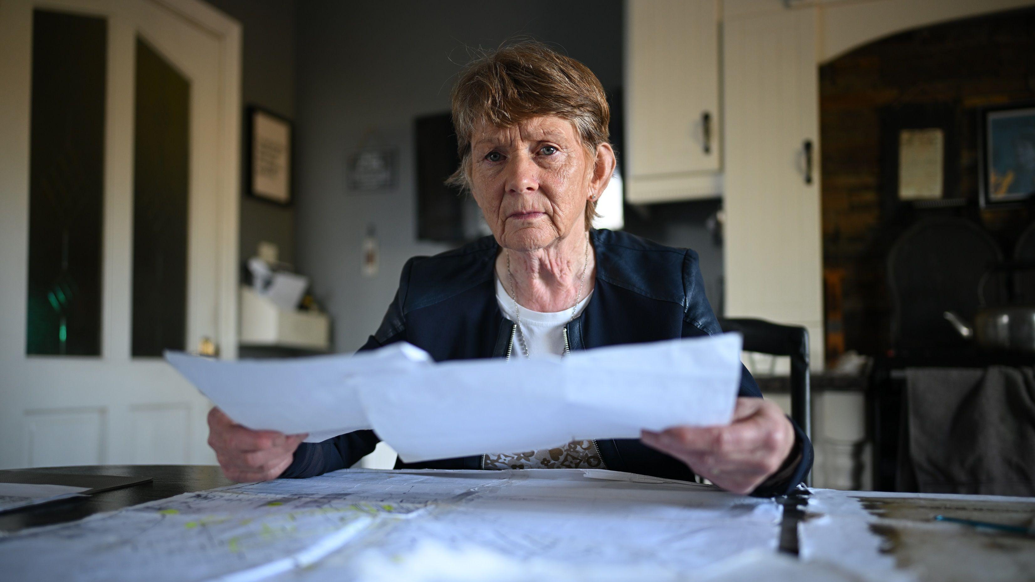 Una mujer mayor está sentada a la mesa de la cocina. Sostiene una hoja de papel en cada mano. Sobre la mesa, frente a ella, hay más documentos y hojas de papel. Tiene el pelo corto, castaño y canoso, y viste una camiseta blanca y una chaqueta azul. Al fondo, hay armarios de cocina y una puerta de cocina con dos ventanas de cristal.