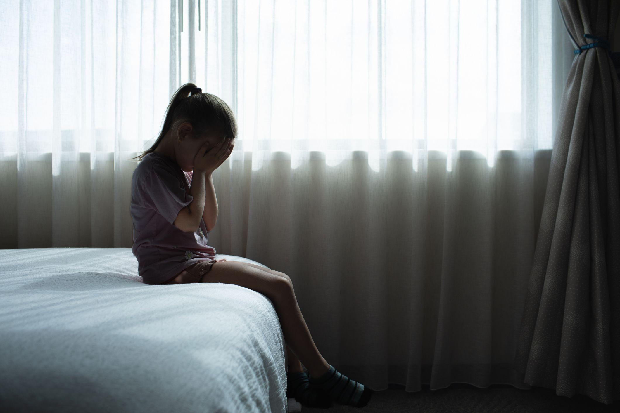Una niña se tapa el rostro mientras está sentada en el borde de una cama. 