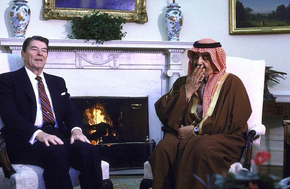 US President Ronald Reagan, wearing a dark navy suit, with white pocket handkerchief, white shirt, red tie with yellow and light-and dark-purple stripes and gold tie-pin and with his hands on his knees, and King Fahd of Saudi Arabia, in traditional dress - a red-and-white keffiyeh headdress and brown-and-gold robes - and covering his mouth with his right hand, on the little finger of which he wears a gold ring with a black stone, as if stifling a yawn, sit beside a roaring fire beneath an ornate mantelpiece topped by a green pot plant and two blue vases decorated in a design featuring yellow flowers, in the White House's Oval Office, in Washington DC, in February 1985. Gold-framed paintings adorn the walls.