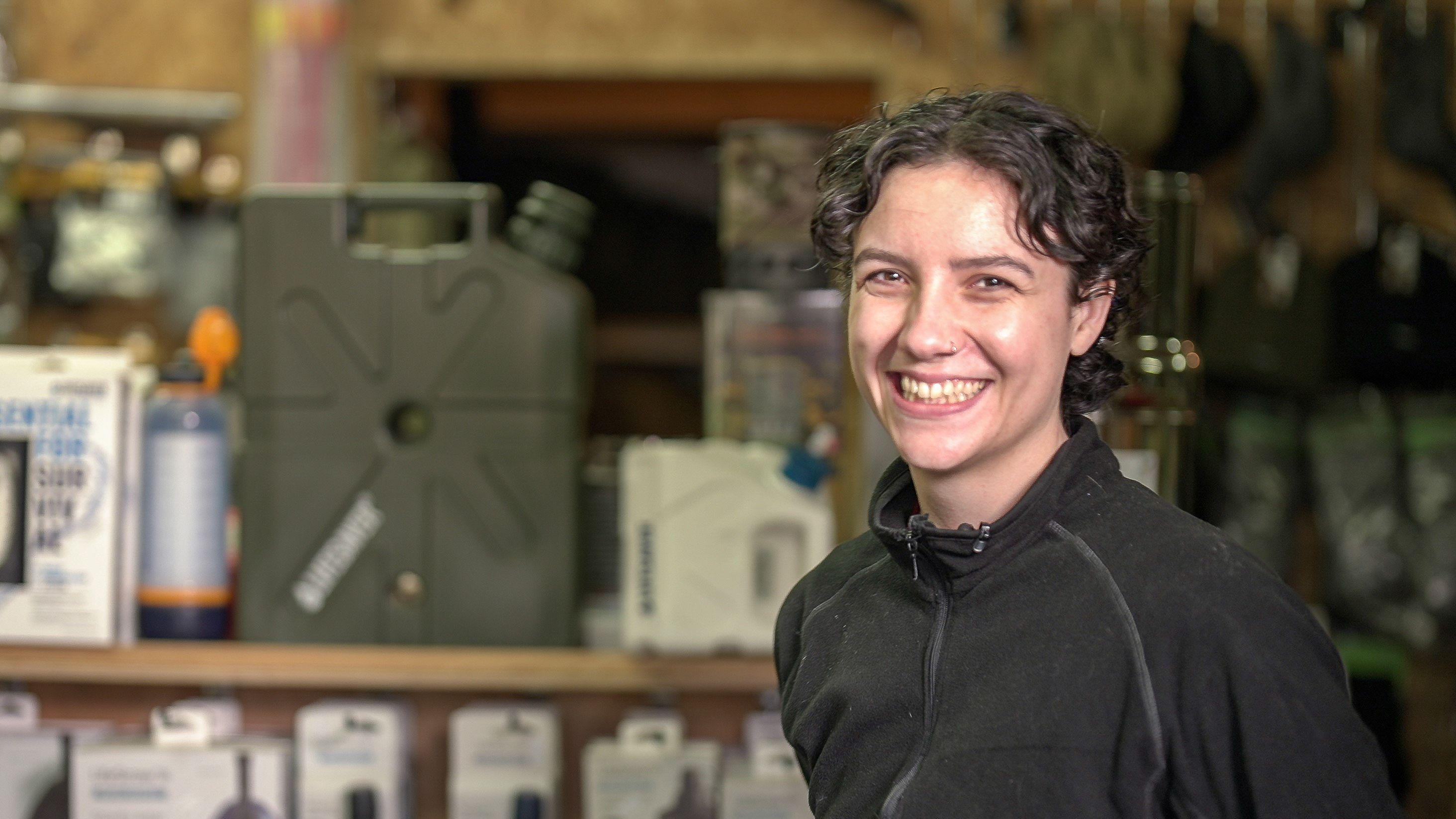 Rowenna &eacute; uma mulher de cabelo castanho curto e cacheado, sorri para a c&acirc;mera. Ela est&aacute; em uma loja cheia de equipamentos de sobreviv&ecirc;ncia. Ela veste um casaco preto