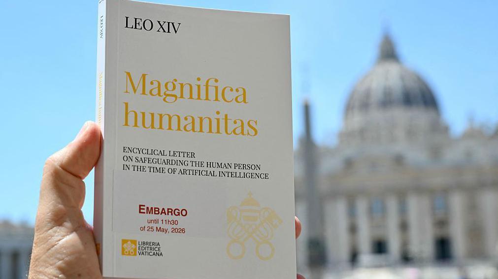 Primer plano de una mano sosteniendo un ejemplar de Magnifica humanitas. En la portada aparece el título de la encíclica, además del embargo que la afecta, en letras rojas. Al fondo se aprecia un cielo azul y lo que parece ser la basílica de San Pedro en Roma.
