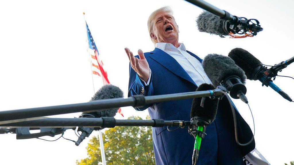 ترامب في صورة مأخوذة من الأسفل إلى الأعلى نهاراً، يرتدي بذلة كحلية رسمية وقميصاً أبيض دون ربطة عنق، وخلفه العلم الأمريكي وأشجار خضراء وأمامه ميكروفونات عدد من وسائل الإعلام.