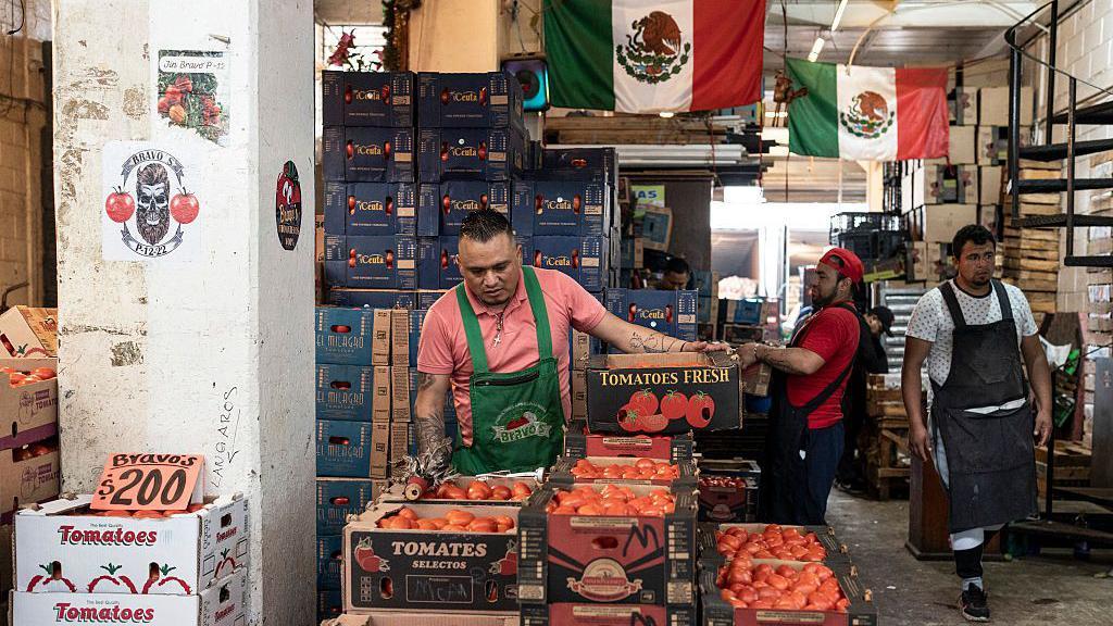 Un vendedor de tomates con la bandera de México al fondo.