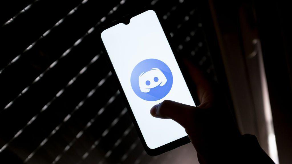 M&atilde;o segurando um celular com um logotipo do Discord na tela, em um ambiente escuro 