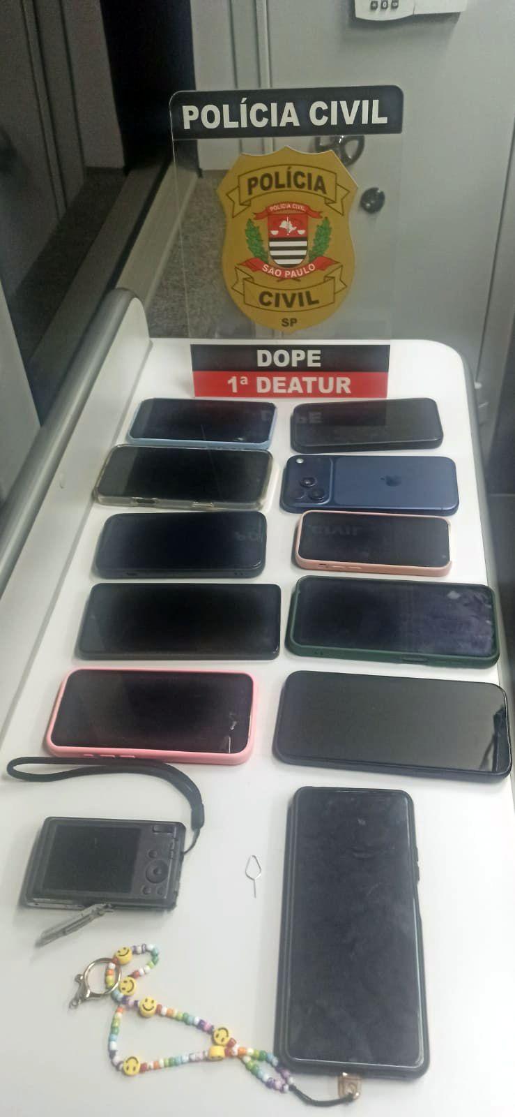 Foto de uma mesa branca com diversos celulares apreendidos dispostos em fileiras, a maioria com a tela apagada e alguns com capas coloridas. Ao fundo, h&aacute; um suporte transparente com o bras&atilde;o e o texto &ldquo;Pol&iacute;cia Civil &ndash; S&atilde;o Paulo&rdquo;, al&eacute;m da identifica&ccedil;&atilde;o &ldquo;DOPE &ndash; 1&ordf; DEATUR&rdquo;. Na parte inferior da imagem, aparecem outros objetos, como uma c&acirc;mera compacta com al&ccedil;a, uma chavinha de remo&ccedil;&atilde;o de chip e um chaveiro com mi&ccedil;angas coloridas e emojis sorridentes. O enquadramento &eacute; vertical e feito de cima para baixo.