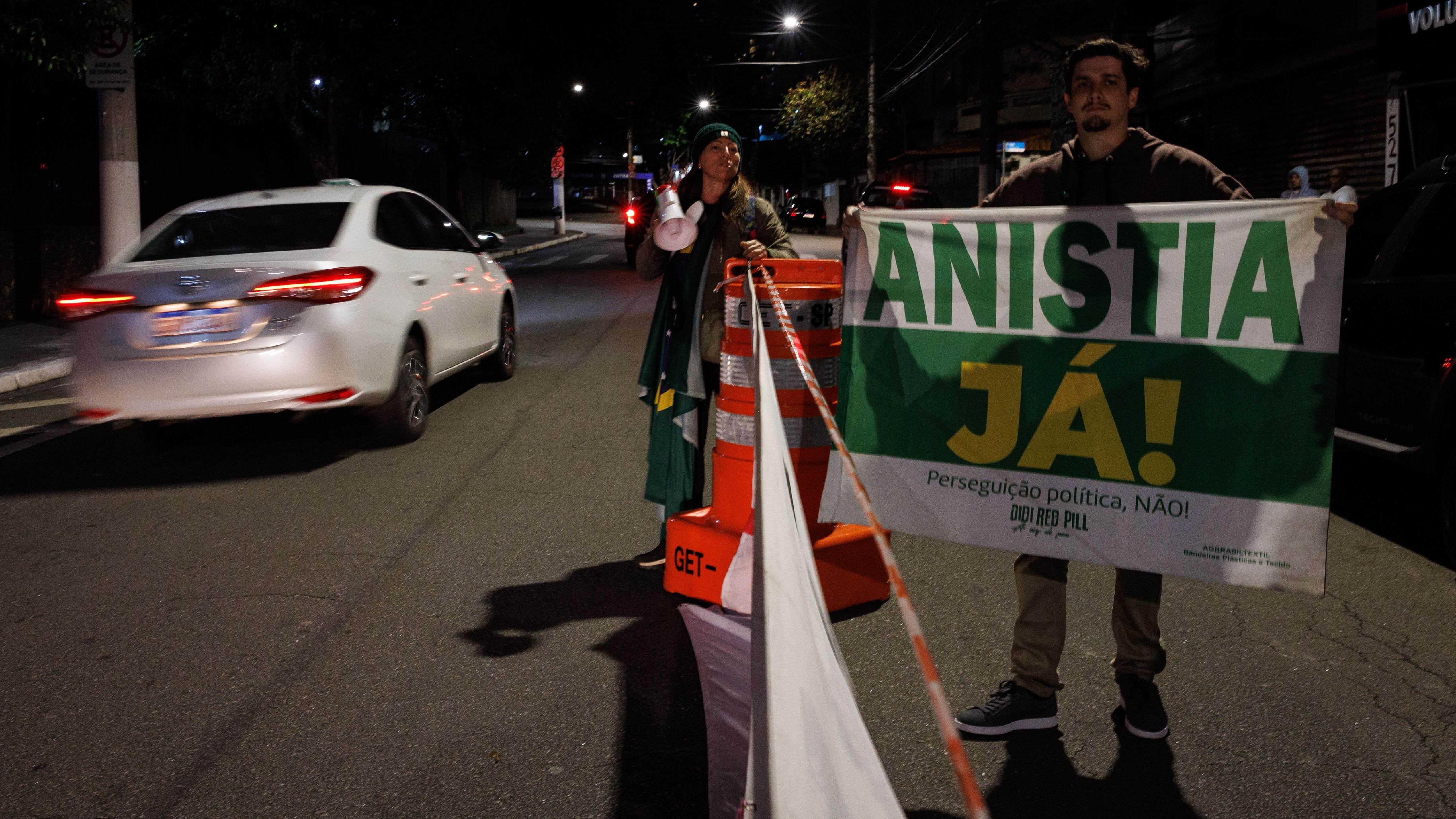 Um homem segura uma faixa escrito 'Anistia já' em um rua, enquanto um táxi passa ao lado. Ao fundo, uma mulher observa com um megafone na mão.