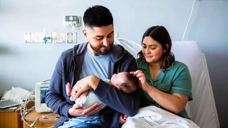 A father and mother sit next to each other in a hospital bed while holding a newborn baby. The father holds the baby in his arms, while the mother strokes its head.