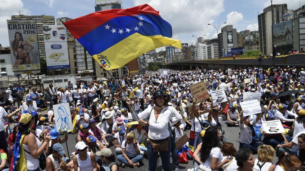 Multid&atilde;o de mulheres, de p&eacute; ou sentadas, com camisetas de cor branca, empunhando bandeiras da Venezuela e carregando cartazes de protesto, em uma das principais ruas de Caracas