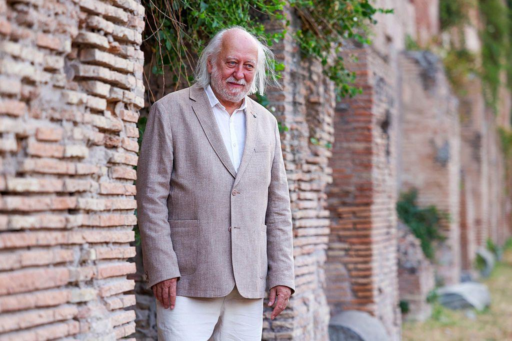 الكاتب كراسنهوركاي رجل ذو شعر أبيض ولحية خفيفة يقف بجانب جدار أثري من الطوب، يرتدي سترة بلون بيج وقميصاً أبيض، ويبدو في خلفية الصورة صف من الجدران القديمة المغطاة بالنباتات.