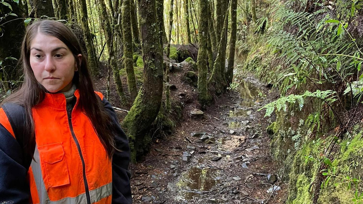 Justine, de pelo largo castaño oscuro, con una chaqueta naranja de alta visibilidad, está de pie en el bosque.