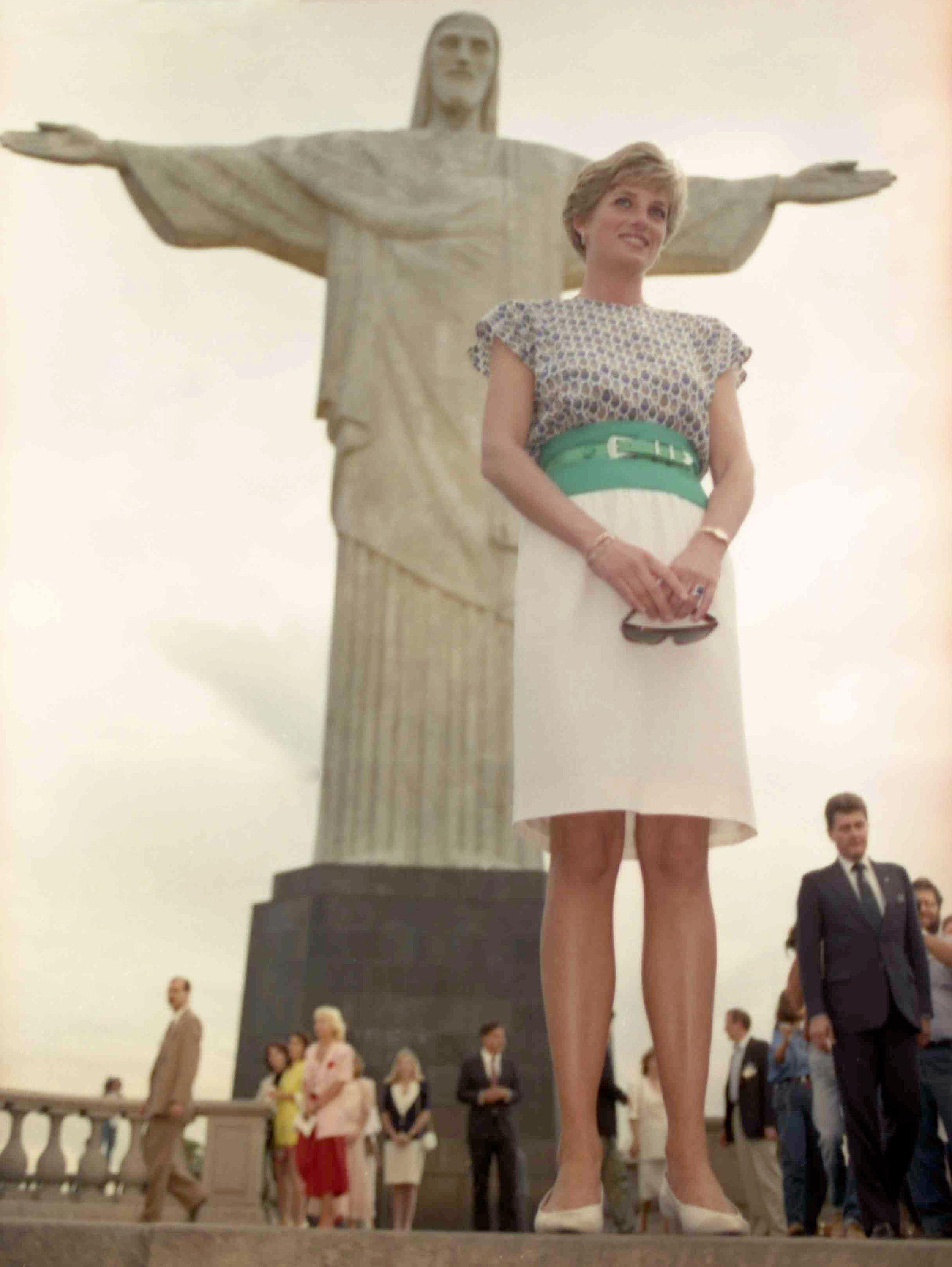 A princesa usa uma saia branca e uma blusa estampada, com um cinco verde. No fundo da foto h&aacute; v&aacute;rias pessoas. 