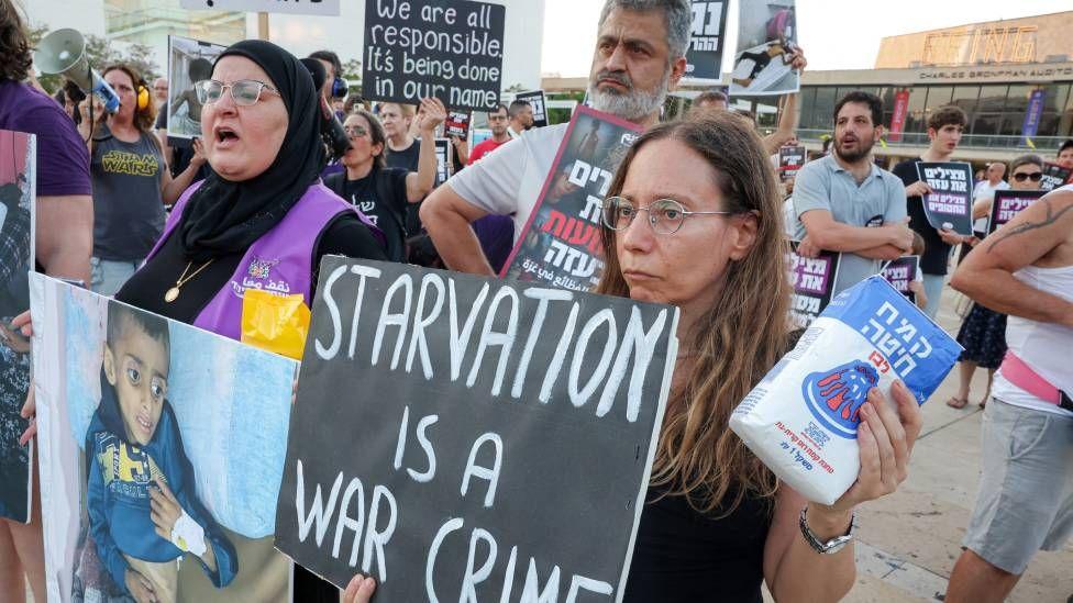 Personas en Tel Aviv con bolsas de comida simbólicas. Llevan pancartas con imágenes de niños en estado de desnutrición y un cartel que dice "la hambruna es un crimen de guerra"
