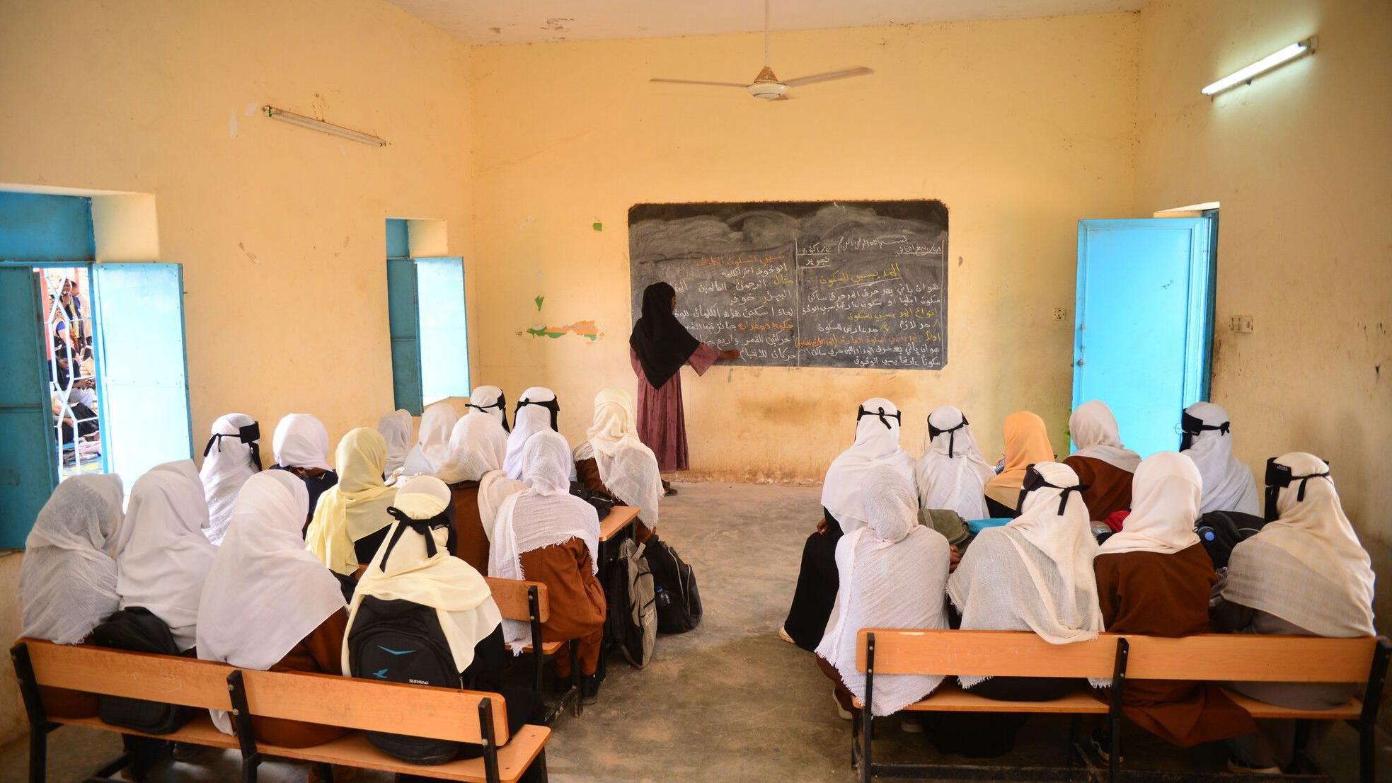 مجموعة من الفتيات تجلس داخل فصل دراسي بمدرسة في ولاية كسلا. وفي مقدمة الفصل، معلمة تكتب دروساً على سبورة وتشرحها.