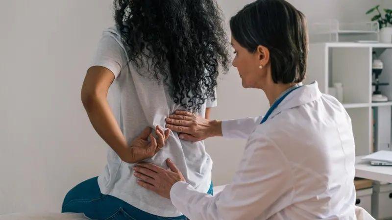 Médica examinando as costas de uma paciente. As duas estão de costas.